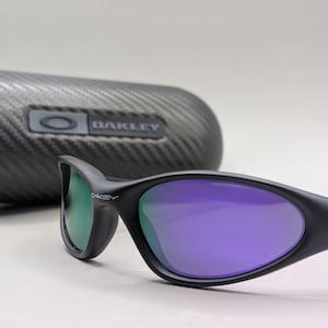 May include: Black Oakley sunglasses with purple lenses and a green tint on the left lens. The sunglasses are set in front of a black, textured case with the Oakley logo. The lenses have a curved shape and the frame is a matte black.