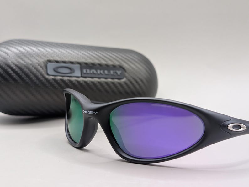 May include: Black Oakley sunglasses with purple lenses and a green tint on the left lens. The sunglasses are set in front of a black, textured case with the Oakley logo. The lenses have a curved shape and the frame is a matte black.