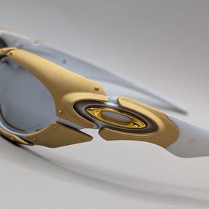May include: Close-up of white and gold-toned sunglasses. The sunglasses feature a gold frame around the lenses and gold accents on the arms. The lenses are a reflective silver color. The design is modern and sporty.