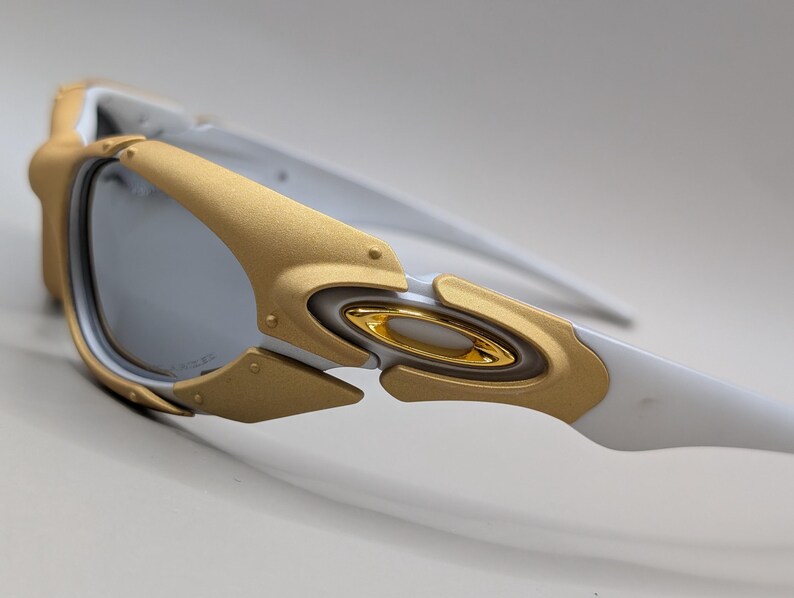 May include: Close-up of white and gold-toned sunglasses. The sunglasses feature a gold frame around the lenses and gold accents on the arms. The lenses are a reflective silver color. The design is modern and sporty.