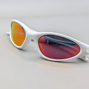 May include: White sunglasses with a streamlined design. The lenses feature a gradient, shifting from orange to red. The frames are bright white, with the Oakley logo visible on the side.