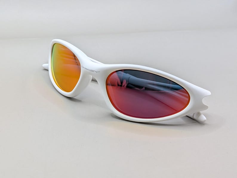 May include: White sunglasses with a streamlined design. The lenses feature a gradient, shifting from orange to red. The frames are bright white, with the Oakley logo visible on the side.