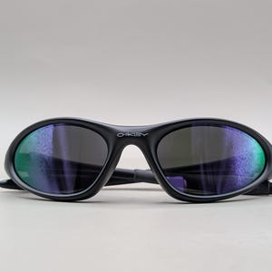 May include: Black Oakley sunglasses with a curved frame and dark lenses. The lenses reflect a mix of green and purple hues. The Oakley logo is visible on the frame.