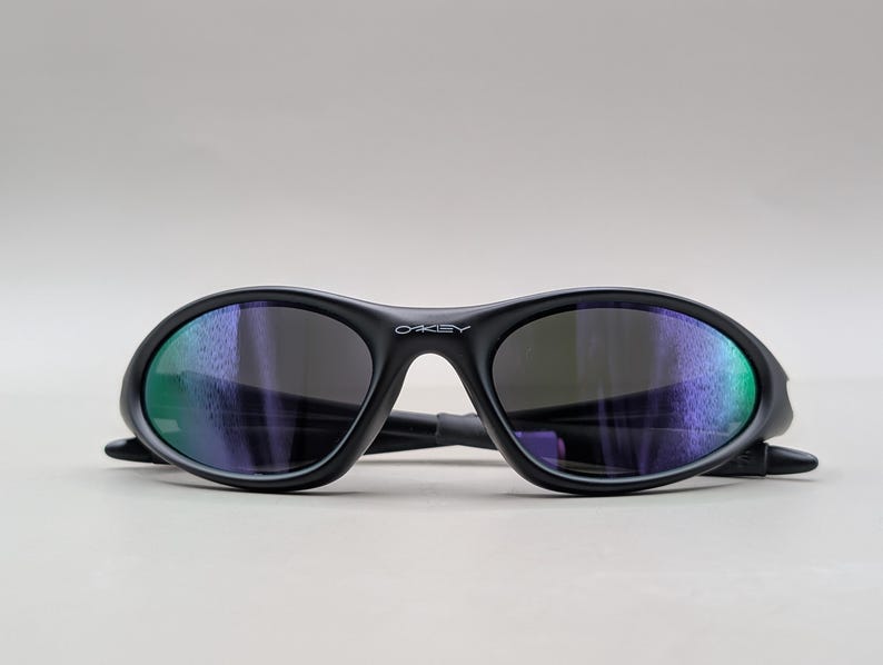 May include: Black Oakley sunglasses with a curved frame and dark lenses. The lenses reflect a mix of green and purple hues. The Oakley logo is visible on the frame.