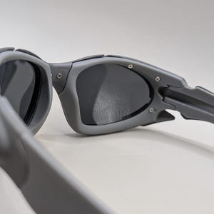 May include: Close-up of gray sunglasses with dark lenses. The sunglasses have a sporty design with a thick frame and arms. The arms are marked "MADE IN U.S.A."