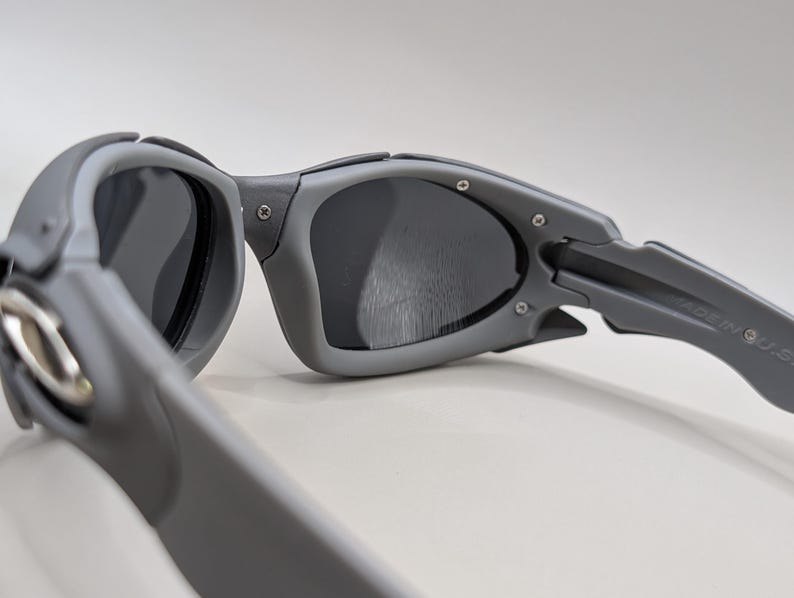 May include: Close-up of gray sunglasses with dark lenses. The sunglasses have a sporty design with a thick frame and arms. The arms are marked "MADE IN U.S.A."