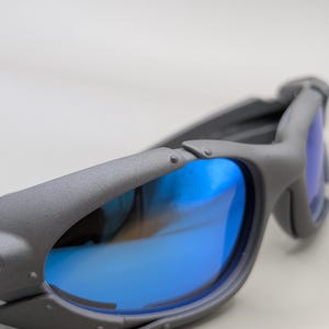 K&ouml;nnte beinhalten: Graue Sonnenbrille mit blau verspiegelten Gl&auml;sern. Die Sonnenbrille hat ein einzigartiges, eckiges Rahmendesign mit Nieten und einer strukturierten Oberfl&auml;che. Die Gl&auml;ser spiegeln eine leuchtend blaue Farbe wider, die mit dem grauen Rahmen kontrastiert.