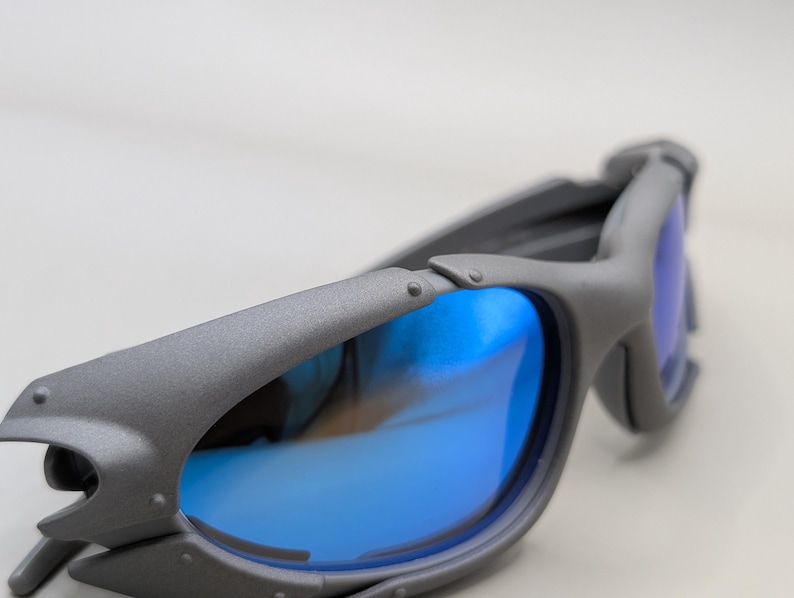K&ouml;nnte beinhalten: Graue Sonnenbrille mit blau verspiegelten Gl&auml;sern. Die Sonnenbrille hat ein einzigartiges, eckiges Rahmendesign mit Nieten und einer strukturierten Oberfl&auml;che. Die Gl&auml;ser spiegeln eine leuchtend blaue Farbe wider, die mit dem grauen Rahmen kontrastiert.