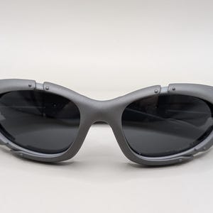 May include: Gray sunglasses with dark lenses. The sunglasses have a unique, futuristic design with a thick frame and riveted details. The lenses are dark, providing a contrast to the gray frame. The arms are thin and extend from the frame.