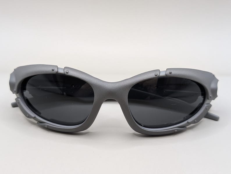 May include: Gray sunglasses with dark lenses. The sunglasses have a unique, futuristic design with a thick frame and riveted details. The lenses are dark, providing a contrast to the gray frame. The arms are thin and extend from the frame.