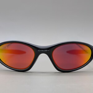 Pode incluir: &Oacute;culos de sol pretos com arma&ccedil;&atilde;o curva e lentes vermelho-alaranjadas. A marca "OAKLEY" est&aacute; impressa em branco na ponte. As lentes t&ecirc;m um efeito gradiente, passando do vermelho para o laranja.