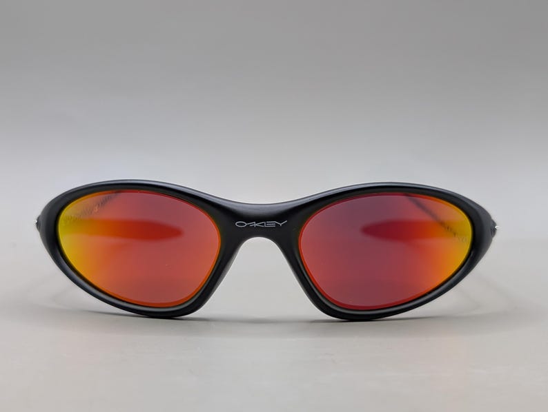 Pode incluir: &Oacute;culos de sol pretos com arma&ccedil;&atilde;o curva e lentes vermelho-alaranjadas. A marca "OAKLEY" est&aacute; impressa em branco na ponte. As lentes t&ecirc;m um efeito gradiente, passando do vermelho para o laranja.