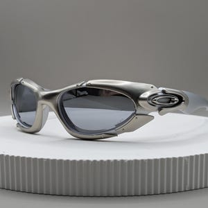 May include: Silver-toned sunglasses with dark lenses. The sunglasses have a futuristic design with a curved frame and a sleek, modern aesthetic. The lenses are a dark gray color, and the arms are a lighter silver.