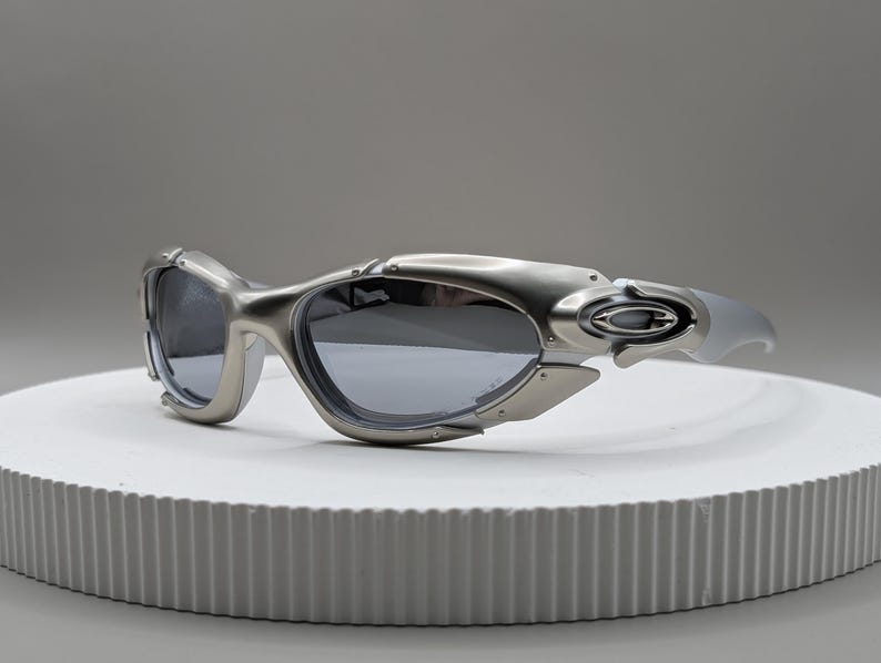 May include: Silver-toned sunglasses with dark lenses. The sunglasses have a futuristic design with a curved frame and a sleek, modern aesthetic. The lenses are a dark gray color, and the arms are a lighter silver.