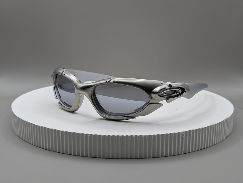 May include: Silver-toned sunglasses with a futuristic design. The sunglasses have a curved frame and dark lenses. The temples are thick and feature a unique design element. The sunglasses are displayed on a white, circular platform.