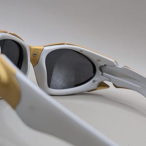 May include: White and gold sunglasses with dark lenses. The sunglasses have a unique design with gold accents on the frame and temples. The lenses are a dark gray color, and the overall style is sporty and modern.