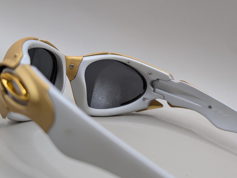 May include: White and gold sunglasses with dark lenses. The sunglasses have a unique design with gold accents on the frame and temples. The lenses are a dark gray color, and the overall style is sporty and modern.