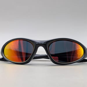 Pode incluir: &Oacute;culos de sol pretos com lentes espelhadas laranja e vermelha. Os &oacute;culos de sol t&ecirc;m uma arma&ccedil;&atilde;o curva e a marca "OAKLEY" &eacute; vis&iacute;vel na ponte.