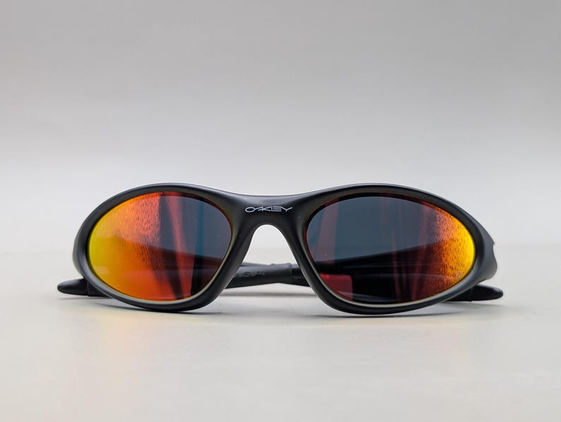 Pode incluir: &Oacute;culos de sol pretos com lentes espelhadas laranja e vermelha. Os &oacute;culos de sol t&ecirc;m uma arma&ccedil;&atilde;o curva e a marca "OAKLEY" &eacute; vis&iacute;vel na ponte.