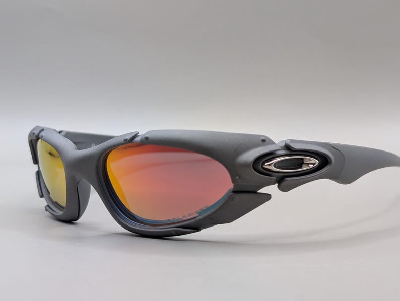 Puede incluir: Gafas de sol grises con lentes naranjas y amarillas. Las gafas tienen un dise&ntilde;o deportivo con una montura gris oscuro y el logotipo de Oakley en el lateral.