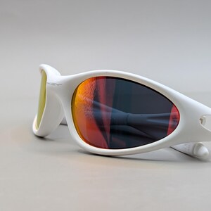 May include: White sunglasses with a curved frame and orange and yellow lenses. The sunglasses are lying on a light grey surface. The lenses reflect the surrounding environment.