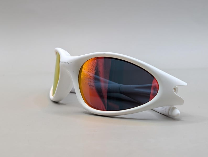 May include: White sunglasses with a curved frame and orange and yellow lenses. The sunglasses are lying on a light grey surface. The lenses reflect the surrounding environment.