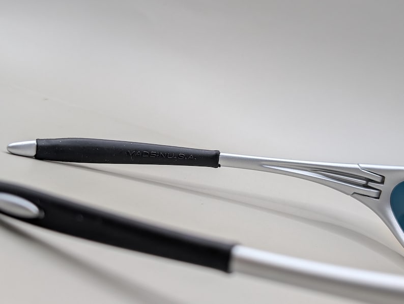 May include: Close-up of silver-framed sunglasses with black rubber grips. The text "MADE IN U.S.A." is visible on the temple arm. The lenses are a light blue color.
