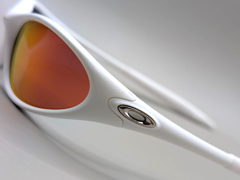 May include: Close-up of white sunglasses with orange-tinted lenses. The sunglasses have a sleek, modern design with a silver accent near the hinge. The lenses reflect a warm, reddish-orange hue.