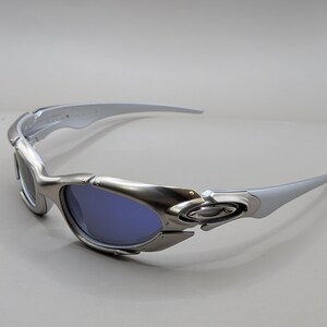 May include: Silver sunglasses with blue lenses. The sunglasses have a sleek, modern design with curved arms and a metallic finish. The Oakley logo is visible on the frame.