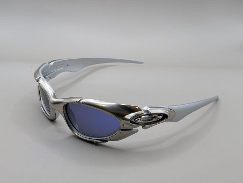 May include: Silver sunglasses with blue lenses. The sunglasses have a sleek, modern design with curved arms and a metallic finish. The Oakley logo is visible on the frame.
