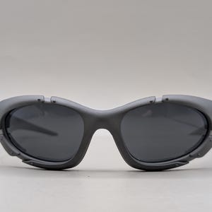 May include: Gray sunglasses with dark lenses. The sunglasses have a curved frame and a unique design with small, decorative details near the temples. The lenses are a dark gray color, providing a sleek and modern look.