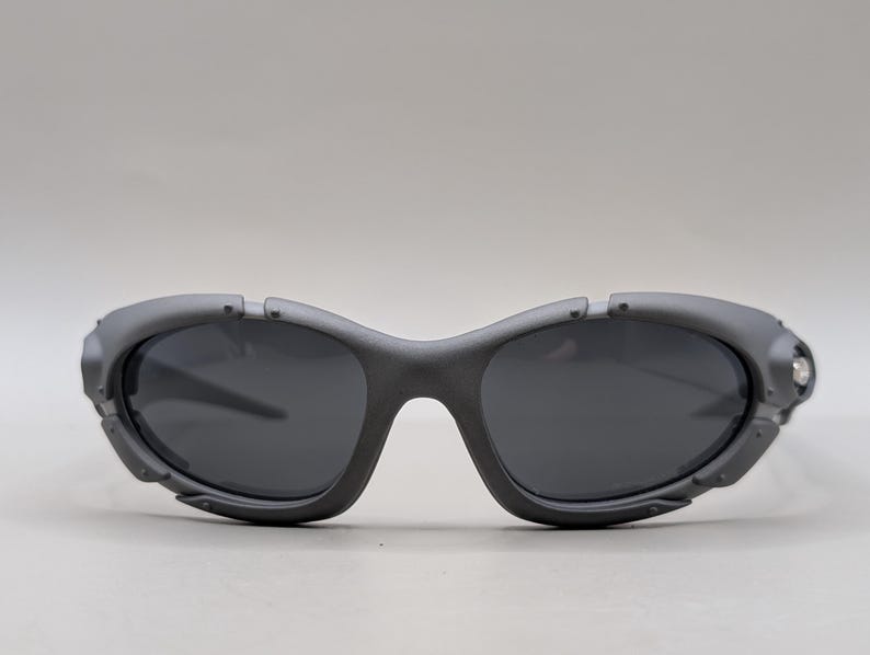 May include: Gray sunglasses with dark lenses. The sunglasses have a curved frame and a unique design with small, decorative details near the temples. The lenses are a dark gray color, providing a sleek and modern look.