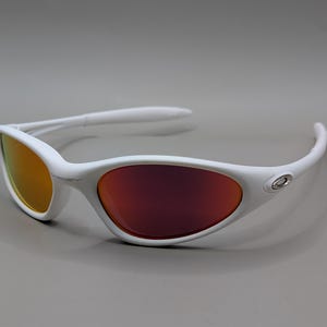 May include: White sunglasses with a curved frame and orange and red lenses. The sunglasses have a sleek, modern design, with a small silver emblem on the side of the frame. The lenses have a gradient effect, transitioning from orange to red.