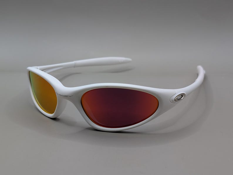 May include: White sunglasses with a curved frame and orange and red lenses. The sunglasses have a sleek, modern design, with a small silver emblem on the side of the frame. The lenses have a gradient effect, transitioning from orange to red.