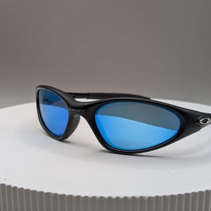 K&ouml;nnte beinhalten: Schwarze Sonnenbrille mit blau verspiegelten Gl&auml;sern. Die Sonnenbrille hat ein schlankes, geschwungenes Design mit dem Markennamen "Oakley" auf dem Rahmen. Die Gl&auml;ser reflektieren eine leuchtend blaue Farbe.