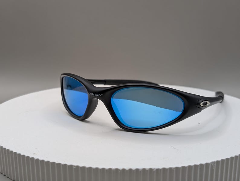 K&ouml;nnte beinhalten: Schwarze Sonnenbrille mit blau verspiegelten Gl&auml;sern. Die Sonnenbrille hat ein schlankes, geschwungenes Design mit dem Markennamen "Oakley" auf dem Rahmen. Die Gl&auml;ser reflektieren eine leuchtend blaue Farbe.