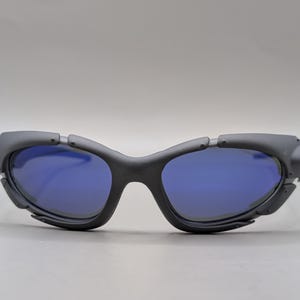 May include: Black sunglasses with blue lenses. The sunglasses have a unique, angular design with a matte finish. The lenses are a vibrant blue, contrasting with the dark frames. The overall style is modern and sporty.