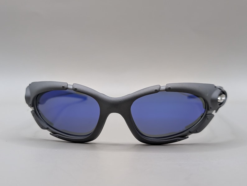 May include: Black sunglasses with blue lenses. The sunglasses have a unique, angular design with a matte finish. The lenses are a vibrant blue, contrasting with the dark frames. The overall style is modern and sporty.