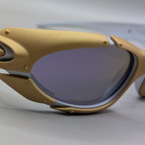 K&ouml;nnte beinhalten: Nahaufnahme einer futuristisch anmutenden Sonnenbrille. Die Fassung ist wei&szlig; und goldfarben, mit einem einzigartigen, kantigen Design. Die Gl&auml;ser sind in einem ged&auml;mpften Lila gehalten, und die Gesamt&auml;sthetik ist elegant und modern.