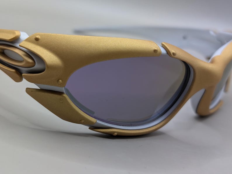 K&ouml;nnte beinhalten: Nahaufnahme einer futuristisch anmutenden Sonnenbrille. Die Fassung ist wei&szlig; und goldfarben, mit einem einzigartigen, kantigen Design. Die Gl&auml;ser sind in einem ged&auml;mpften Lila gehalten, und die Gesamt&auml;sthetik ist elegant und modern.