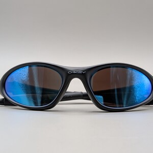 K&ouml;nnte beinhalten: Schwarze Oakley-Sonnenbrille mit blau verspiegelten Gl&auml;sern. Die Sonnenbrille hat einen geschwungenen Rahmen und das Oakley-Logo ist auf dem B&uuml;gel sichtbar. Die Gl&auml;ser sind braun get&ouml;nt.
