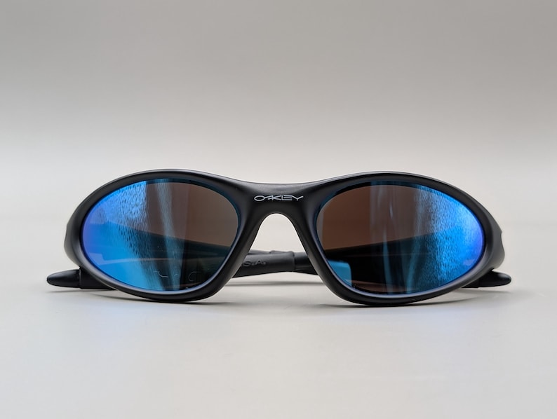 K&ouml;nnte beinhalten: Schwarze Oakley-Sonnenbrille mit blau verspiegelten Gl&auml;sern. Die Sonnenbrille hat einen geschwungenen Rahmen und das Oakley-Logo ist auf dem B&uuml;gel sichtbar. Die Gl&auml;ser sind braun get&ouml;nt.