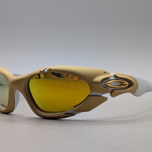 Pu&ograve; includere: Occhiali da sole Oakley con montatura oro e bianca e lenti gialle. Gli occhiali da sole hanno un design futuristico con una forma curva e un logo Oakley prominente sull'asta.
