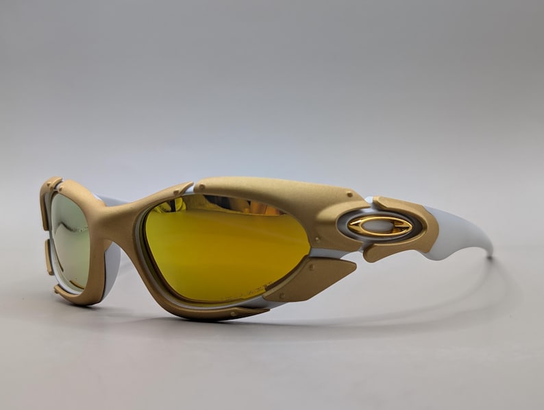 Pu&ograve; includere: Occhiali da sole Oakley con montatura oro e bianca e lenti gialle. Gli occhiali da sole hanno un design futuristico con una forma curva e un logo Oakley prominente sull'asta.