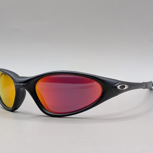 Pode incluir: &Oacute;culos de sol esportivos pretos com arma&ccedil;&atilde;o curva e lentes espelhadas vermelho-alaranjadas. A lente esquerda tem um tom amarelo. O logotipo da Oakley &eacute; vis&iacute;vel na arma&ccedil;&atilde;o. Projetados para esportes e atividades ao ar livre.