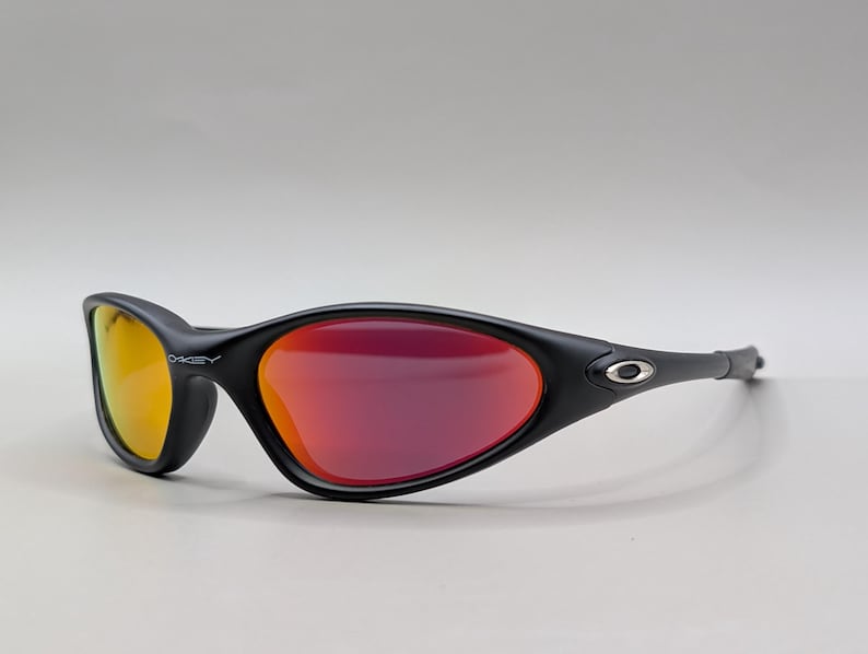 Pode incluir: &Oacute;culos de sol esportivos pretos com arma&ccedil;&atilde;o curva e lentes espelhadas vermelho-alaranjadas. A lente esquerda tem um tom amarelo. O logotipo da Oakley &eacute; vis&iacute;vel na arma&ccedil;&atilde;o. Projetados para esportes e atividades ao ar livre.