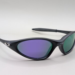 May include: Black sunglasses with purple and green lenses. The sunglasses have a sleek, curved design with the brand name "Oakley" visible on the frame. The lenses reflect light, creating a vibrant color contrast.