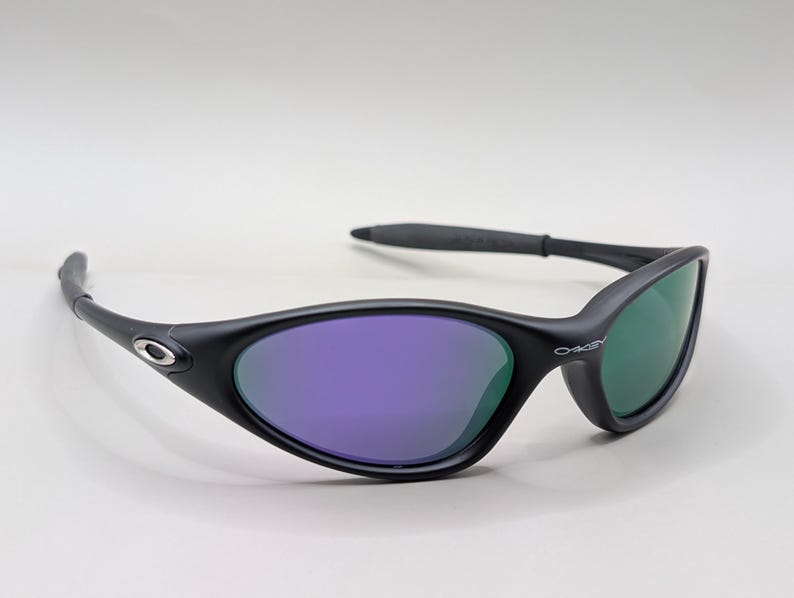 May include: Black sunglasses with purple and green lenses. The sunglasses have a sleek, curved design with the brand name "Oakley" visible on the frame. The lenses reflect light, creating a vibrant color contrast.