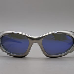 May include: Silver sunglasses with blue lenses. The sunglasses feature a futuristic design with a curved frame and a sleek, metallic finish. The lenses are a deep blue colour, contrasting with the silver frame.