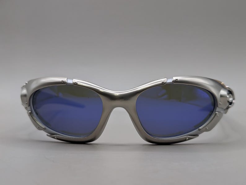 May include: Silver sunglasses with blue lenses. The sunglasses feature a futuristic design with a curved frame and a sleek, metallic finish. The lenses are a deep blue colour, contrasting with the silver frame.